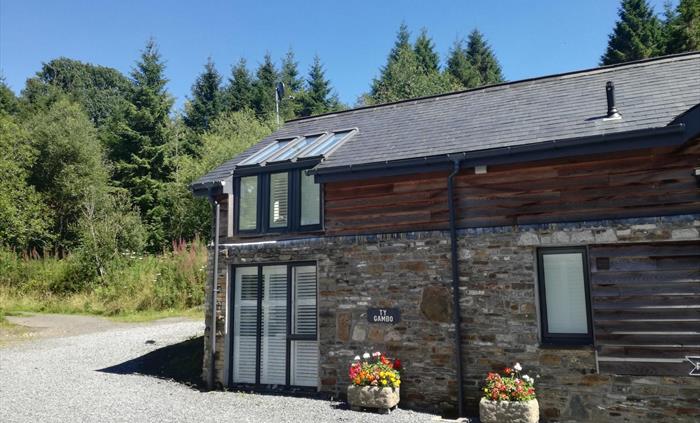 A converted stone barn nestled at the forest’s edge, featuring solar panels, shuttered windows, and vibrant flower displays by the entrance.